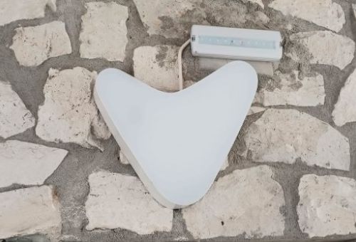 Un dispositivo Visualfy blanco con forma de corazón invertido o "V" estilizada, emitiendo una luz suave, montado sobre una pared de piedras irregulares. Este es un sistema inteligente de detección de sonidos de emergencia. Encima y ligeramente a la derech