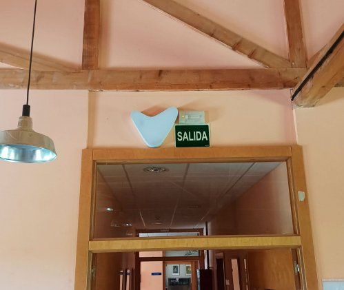 Un dispositivo Visualfy blanco en forma de corazón invertido o "V" estilizada montado sobre una pared de color melocotón pálido, junto a un letrero de "SALIDA" verde con letras blancas. Debajo, se ve la parte superior de un marco de puerta de madera con u