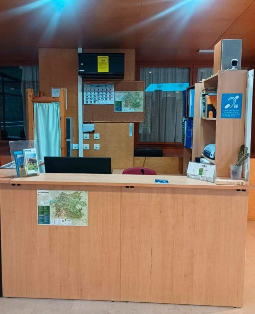 Una recepción de madera clara con un monitor de ordenador negro. Detrás del mostrador, en la pared de madera, hay un corcho con un calendario, un mapa, y un cartel de advertencia amarillo. A la derecha, una estantería de madera contiene libros y un cartel