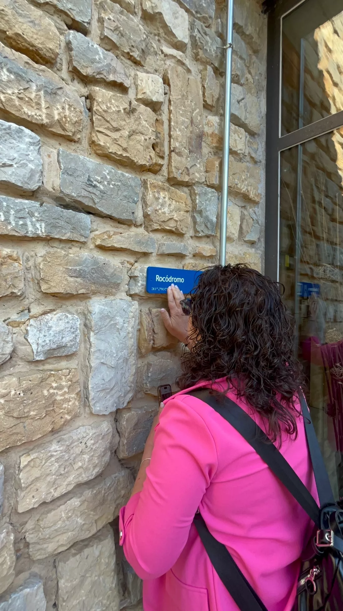 Mujer toca una señal en braille que indica "Rocódromo" en una pared de piedra.